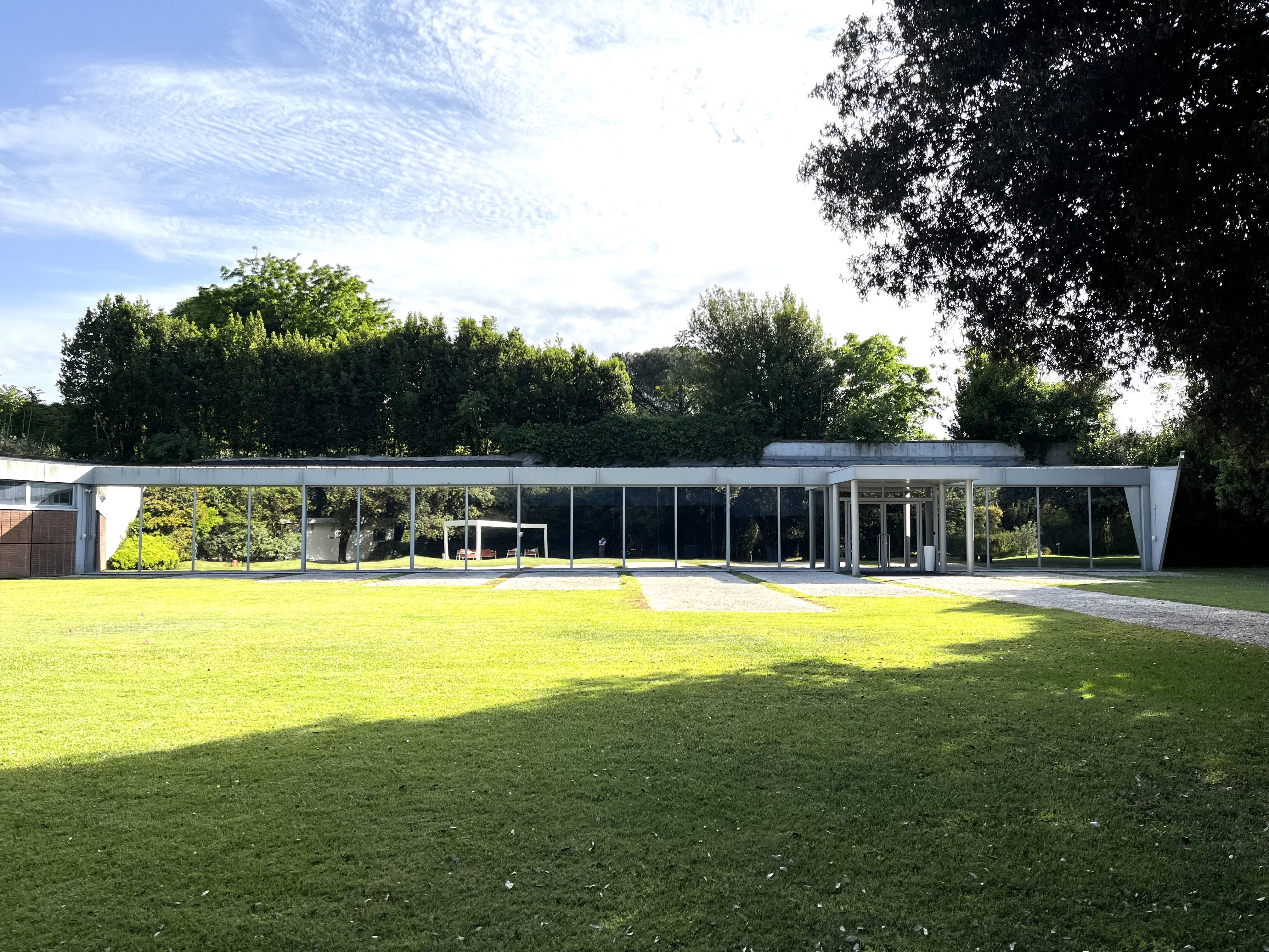 Long glass-fronted pavilion in a sunny park, surrounded by a green lawn and trees.