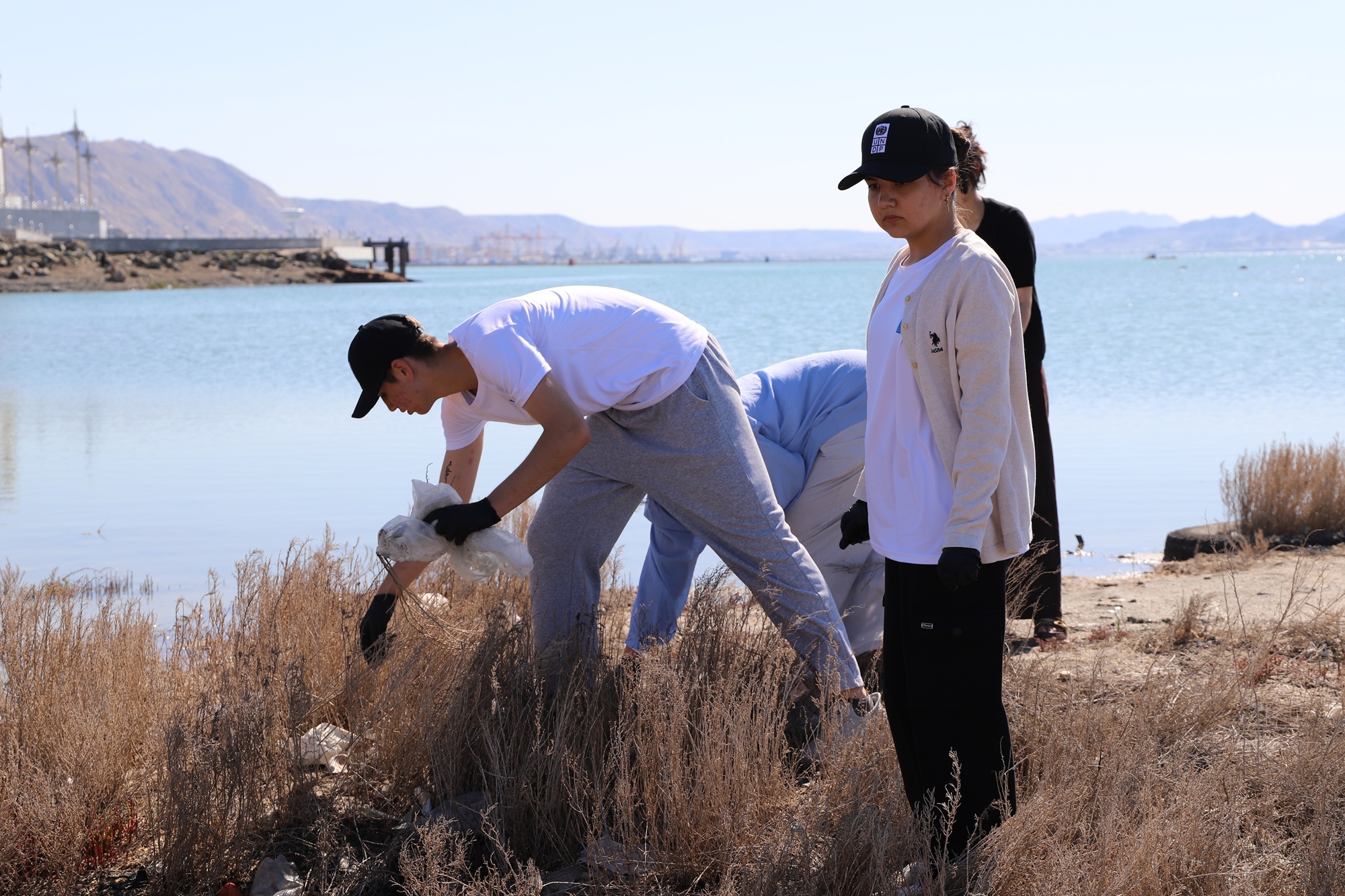 Photograph of three people by a lake, one bending to touch dry grass, mountains in distance.