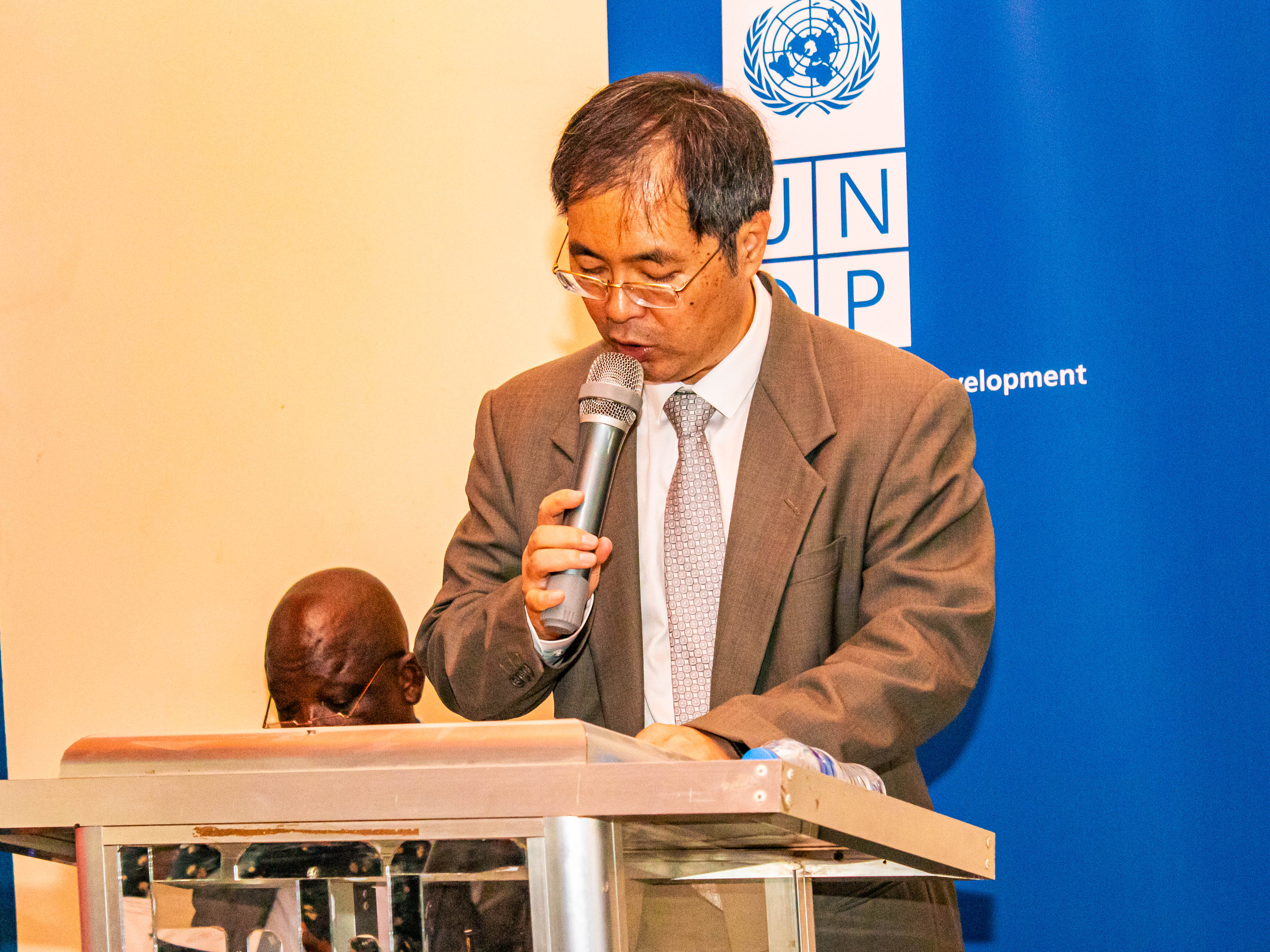 Photograph of a man in a suit speaking at a podium with microphone; blue banner behind.