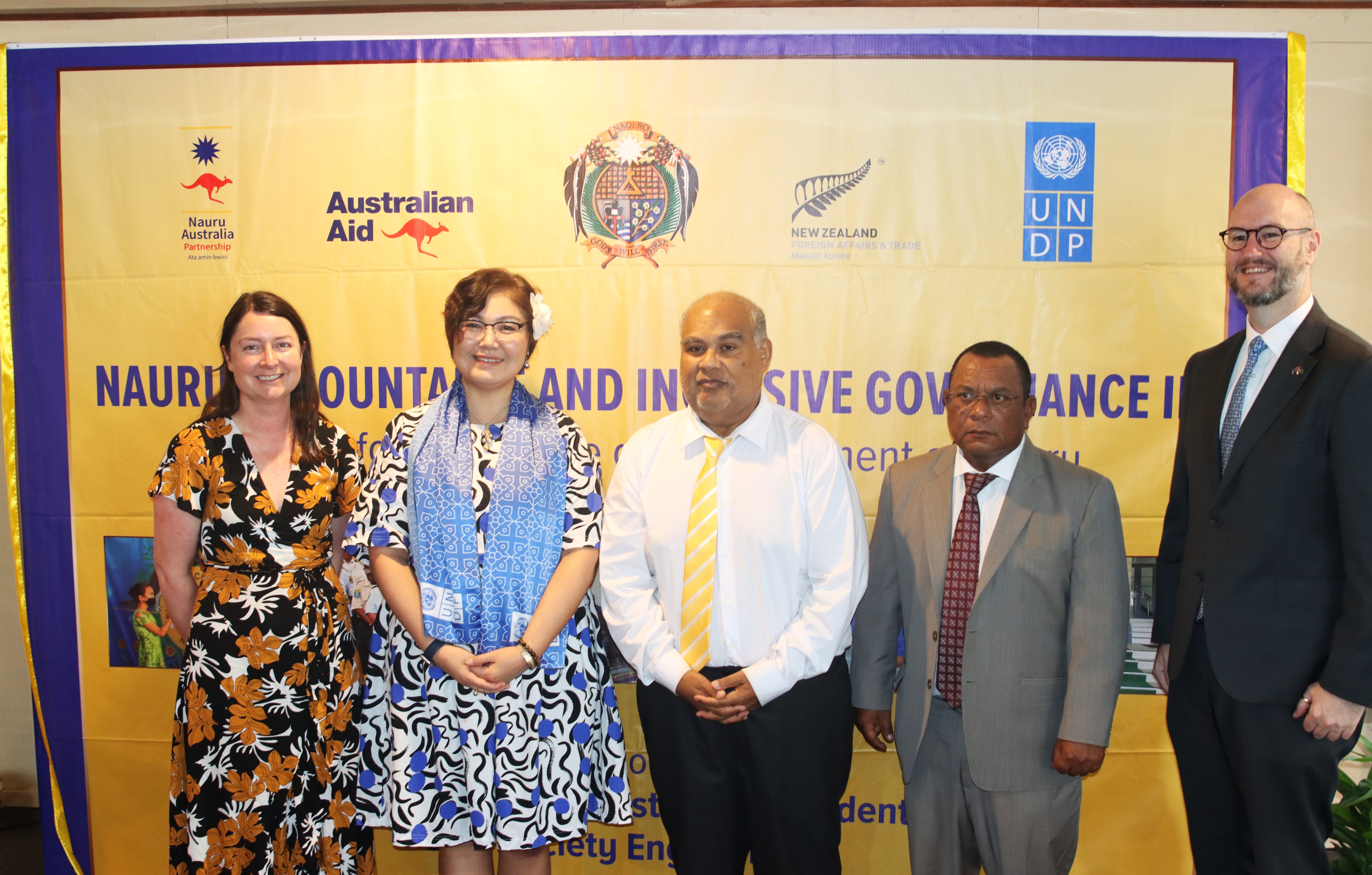 Five people in business attire pose in front of a yellow conference banner with logos.