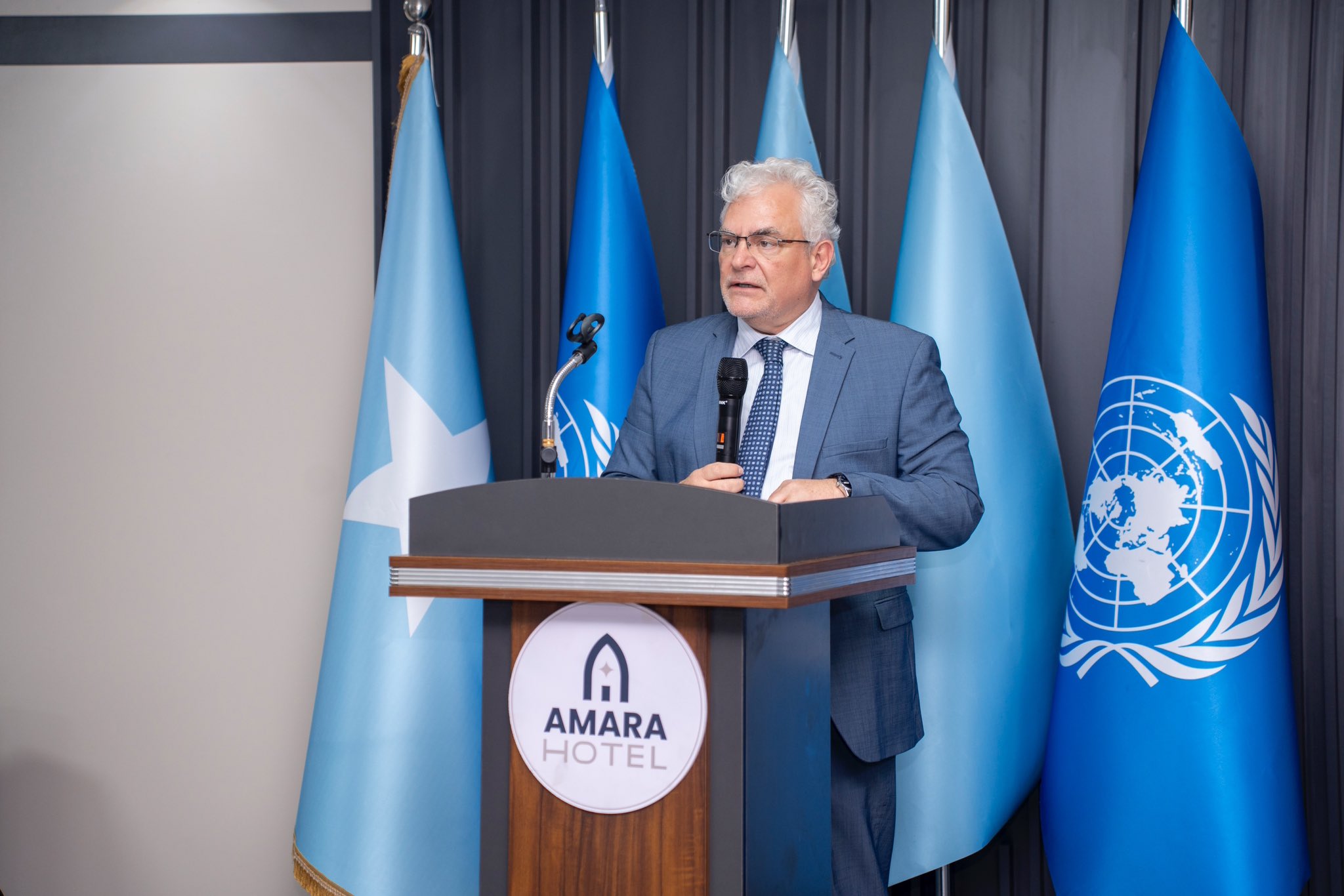 Man in a blue suit with gray hair delivers a speech at a podium, flags behind him.