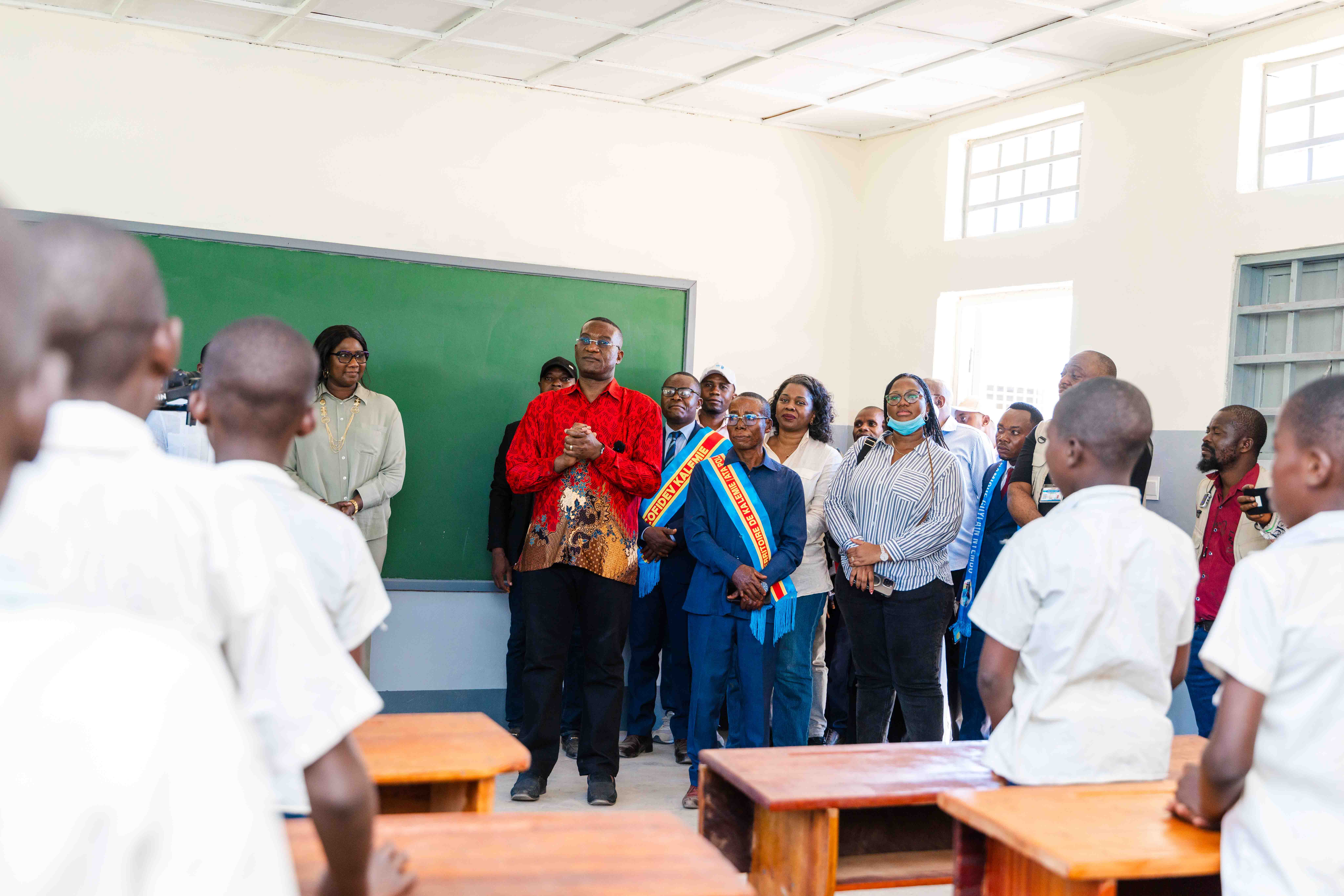Group of people in a classroom stand around desks as a man in red speaks near a green chalkboard.