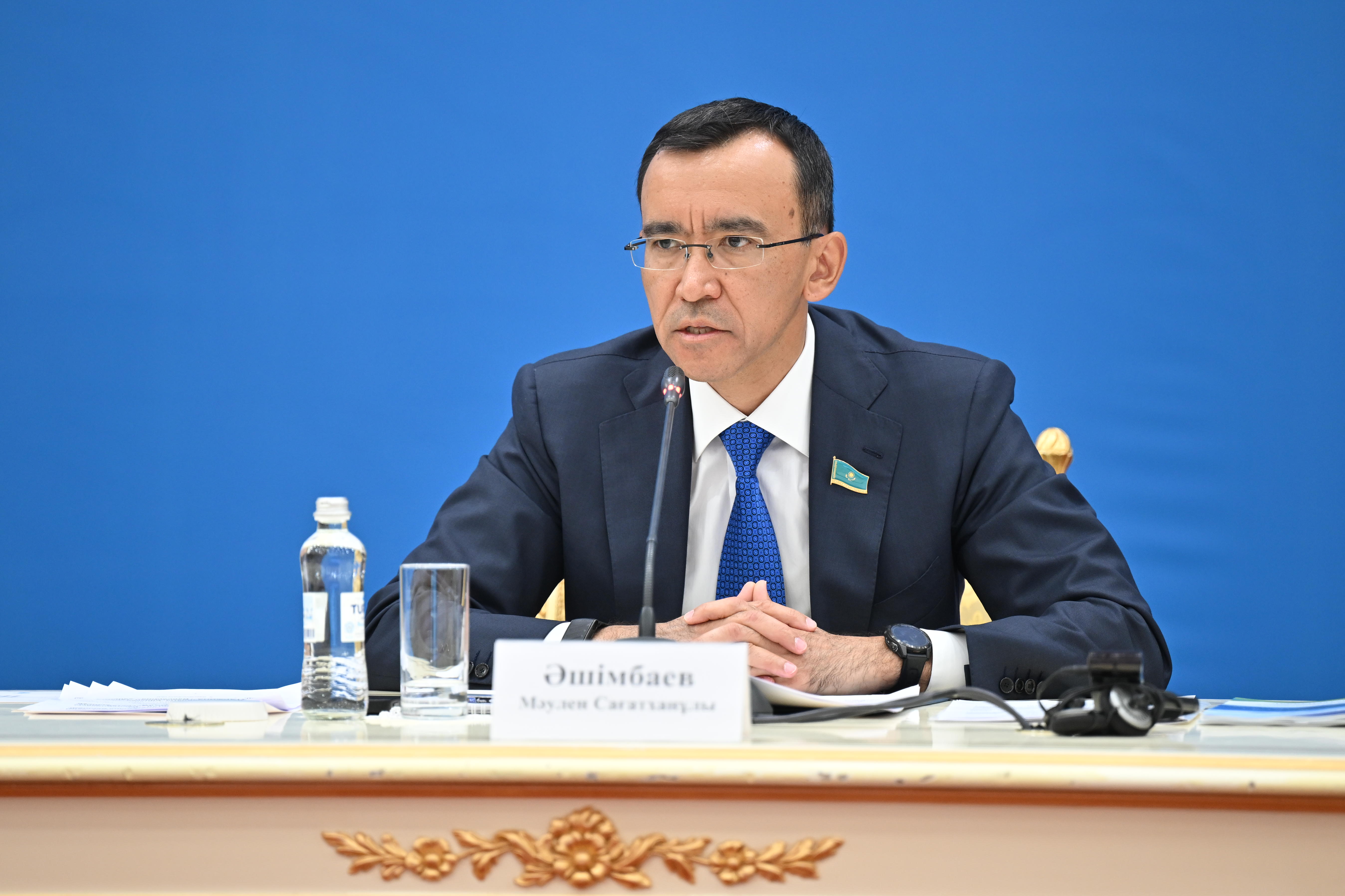 Man in suit seated at a desk on blue backdrop, blurred face, with water bottle and papers.