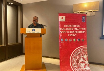 Photograph of a man speaking at a podium beside a red banner in a conference room.