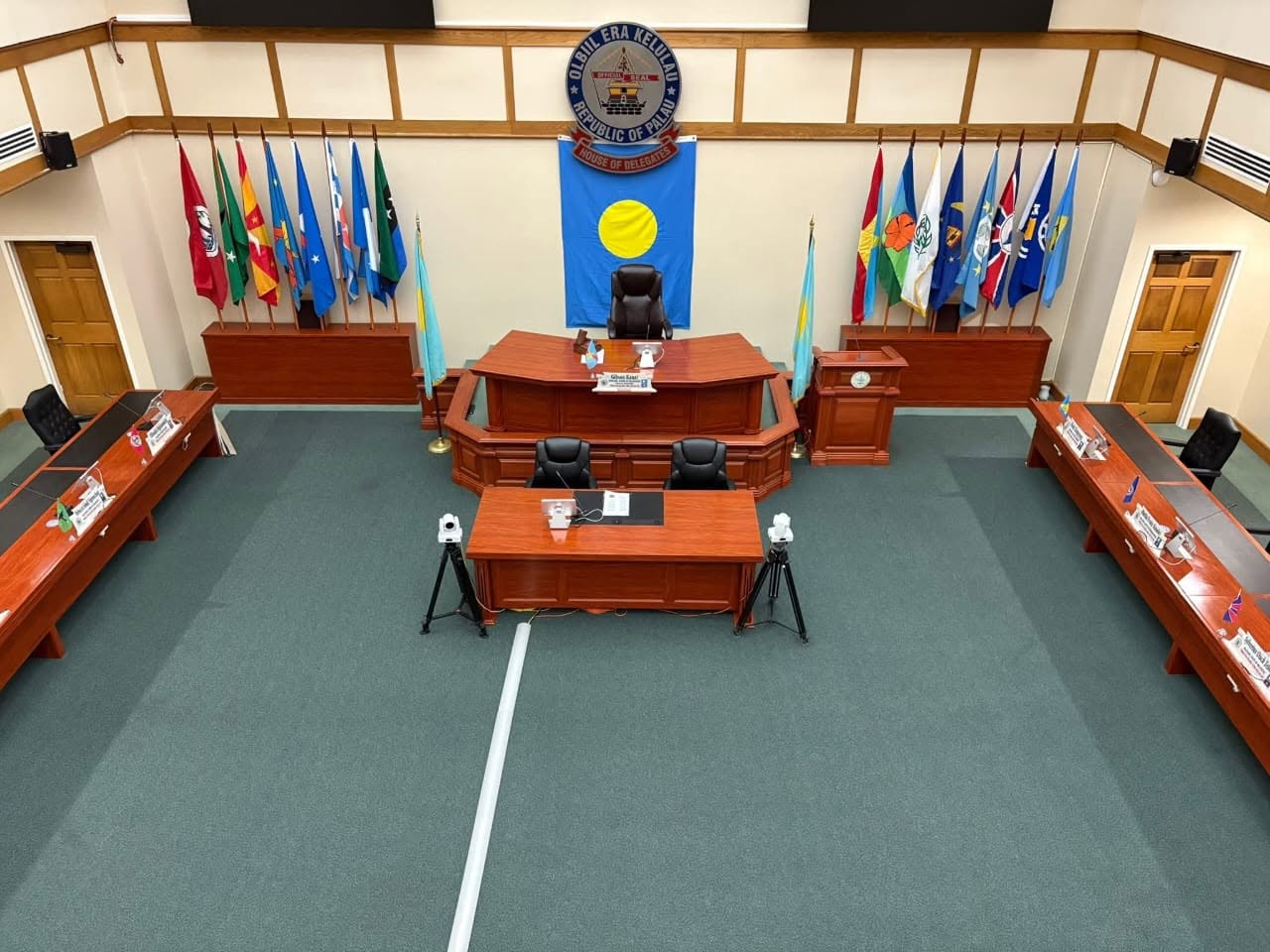 Empty council chamber with blue crest on wall behind a central dais and flags along the sides.