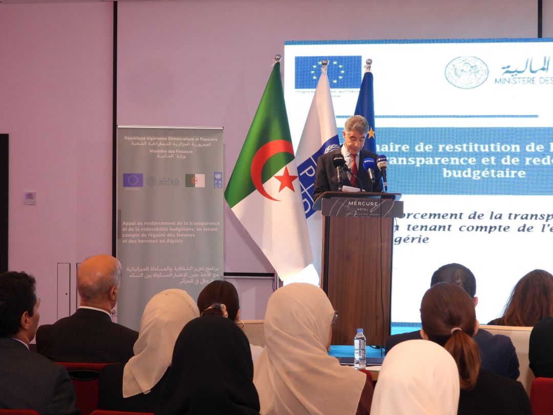 Speaker at podium addressing an audience in a conference room; flags and banners on stage.