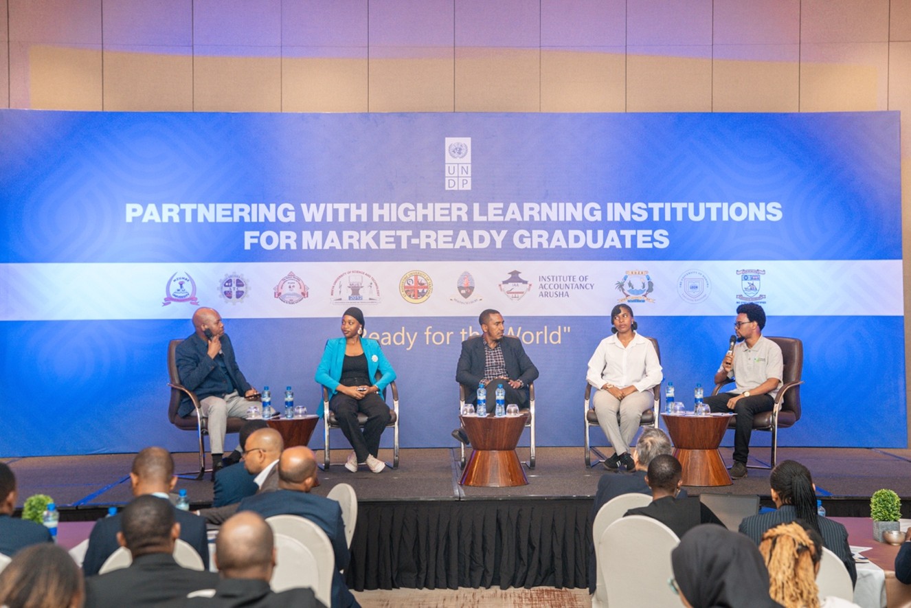Panel discussion on stage with multiple participants; audience seated, blue banner behind.