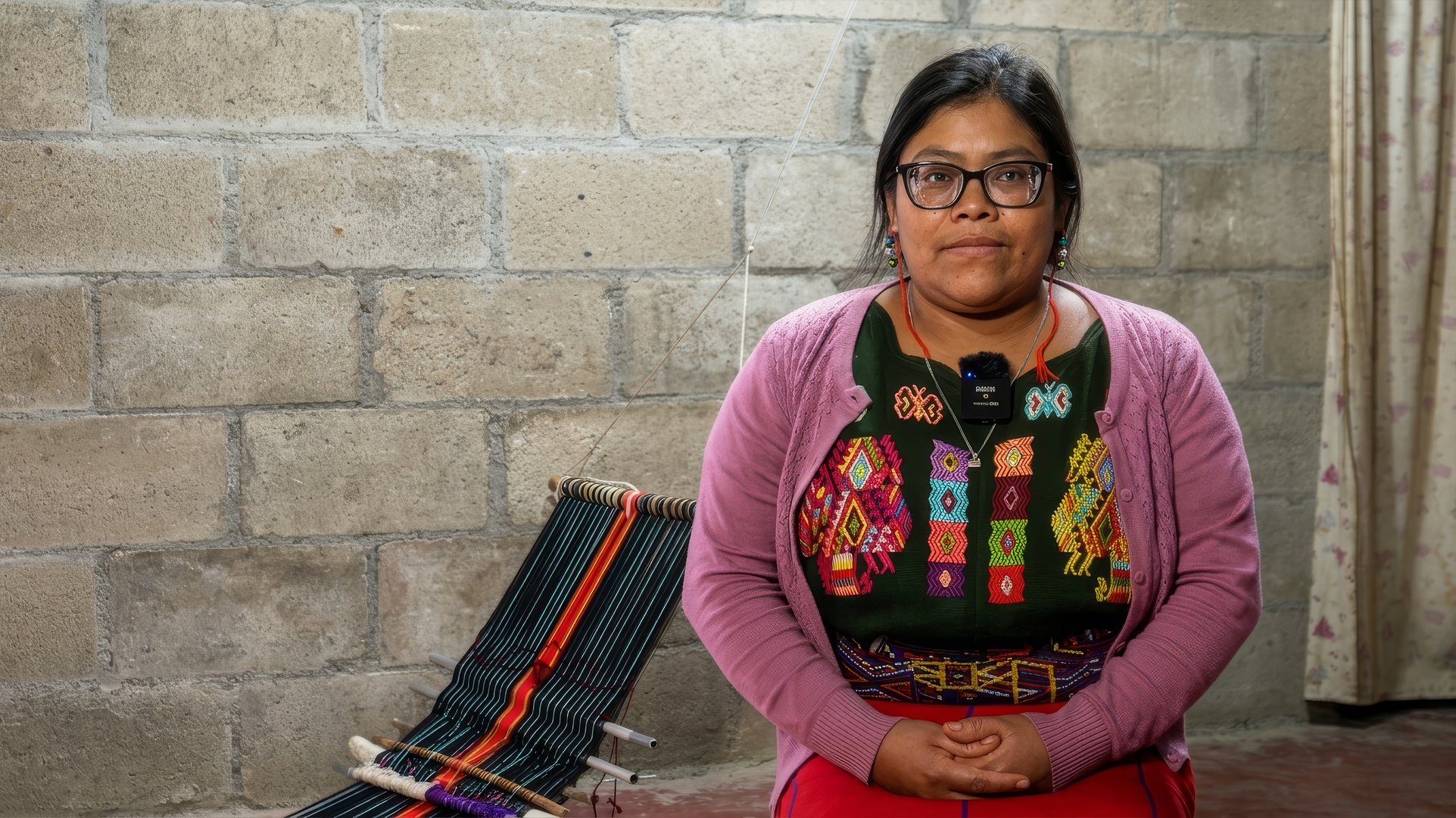 Photo: Person seated against a brick wall, wearing a pink cardigan and colorful embroidered top.