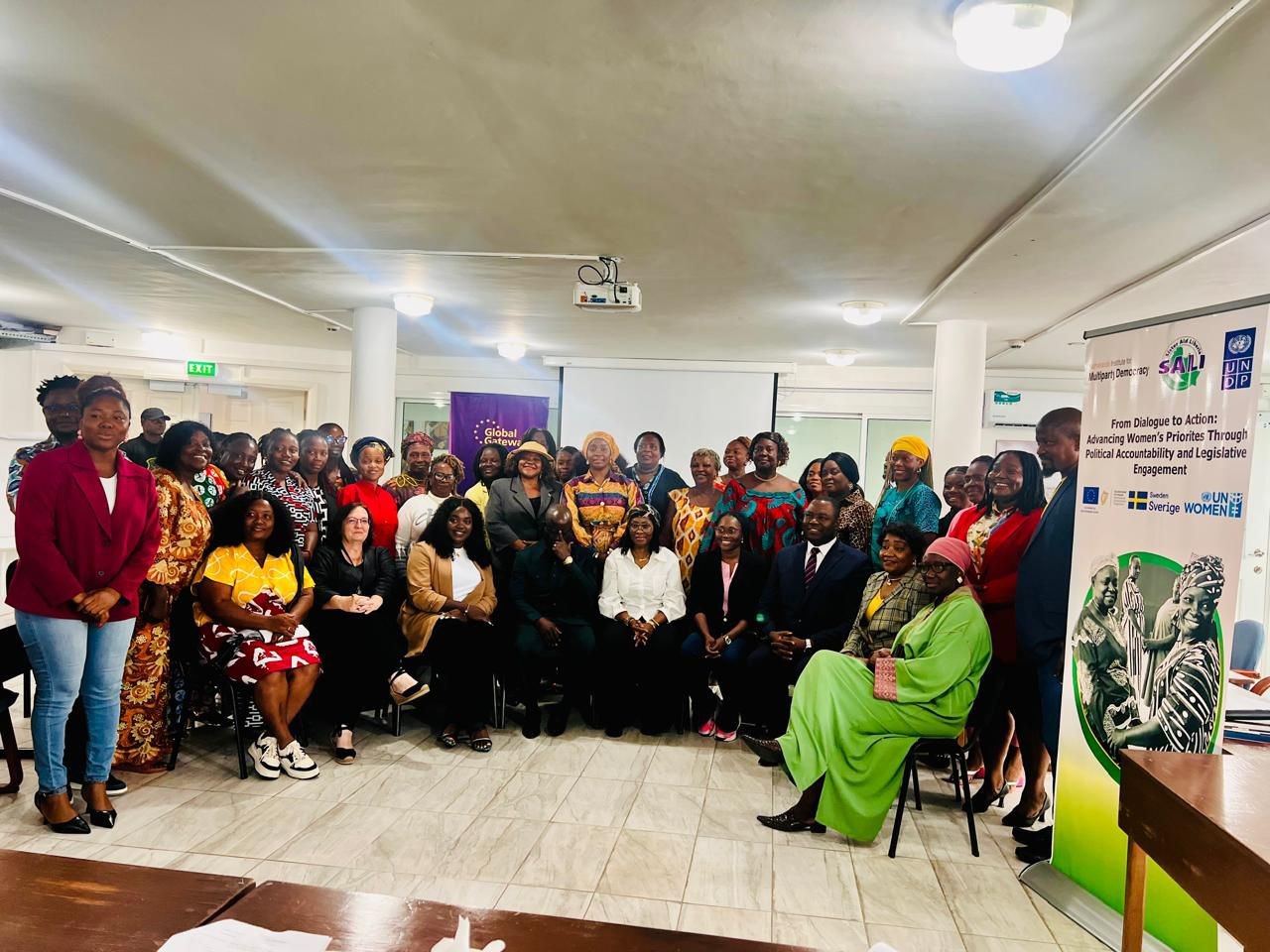 Group photo of many people gathered in a bright community hall, banner on the right.