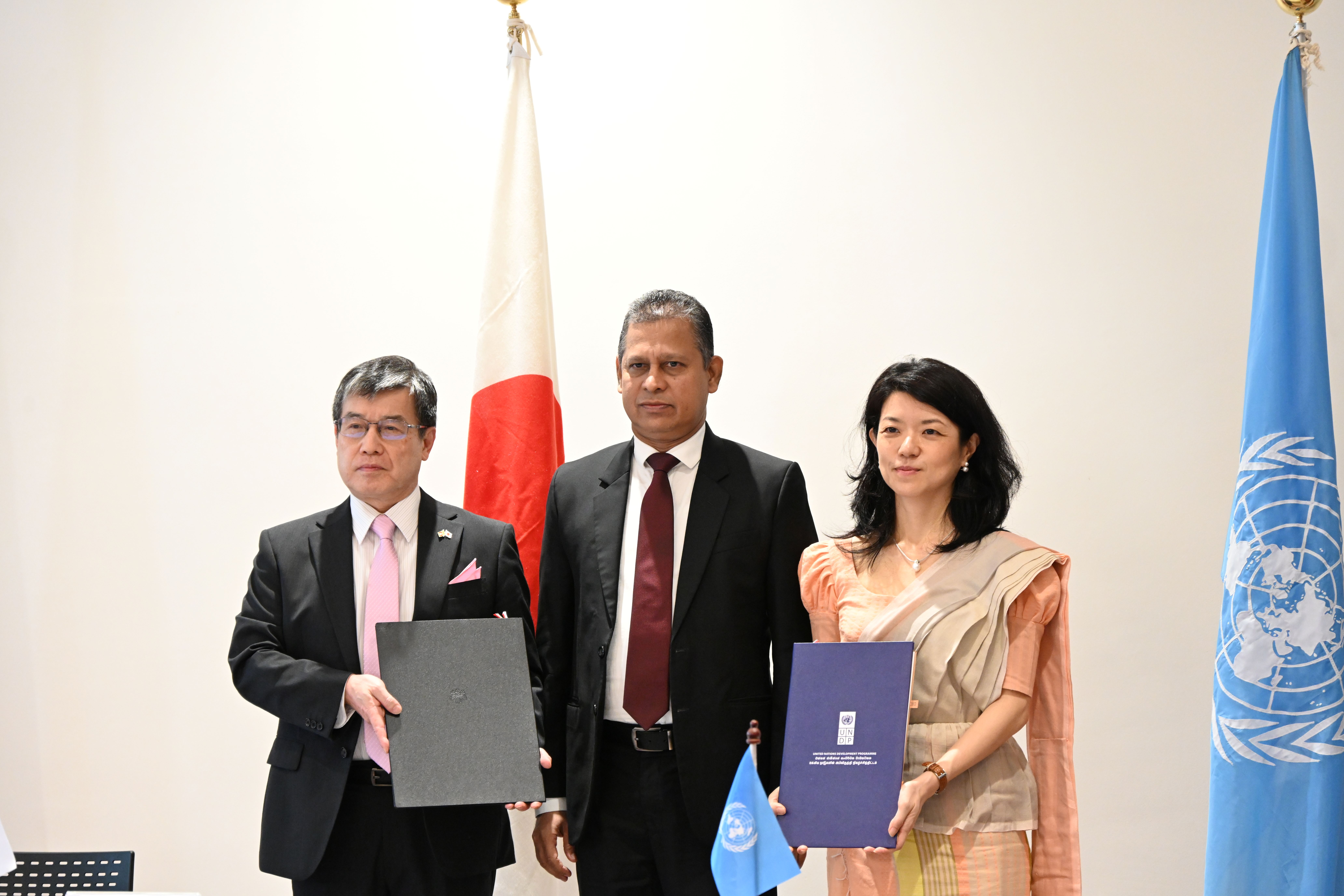 Three officials stand with documents in front of UN and national flags.