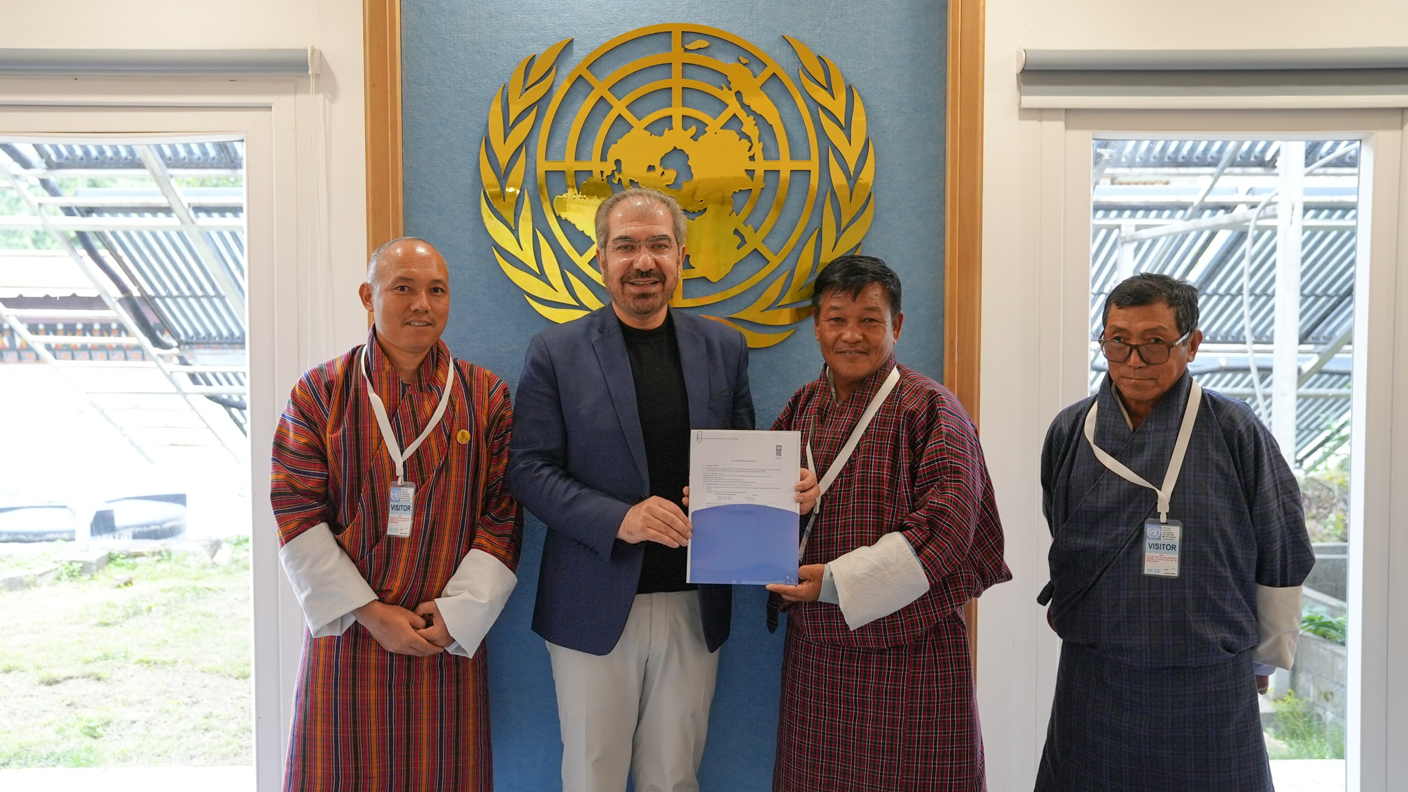 Photo: four people in a meeting room before a UN emblem; central man in blue holds a document.