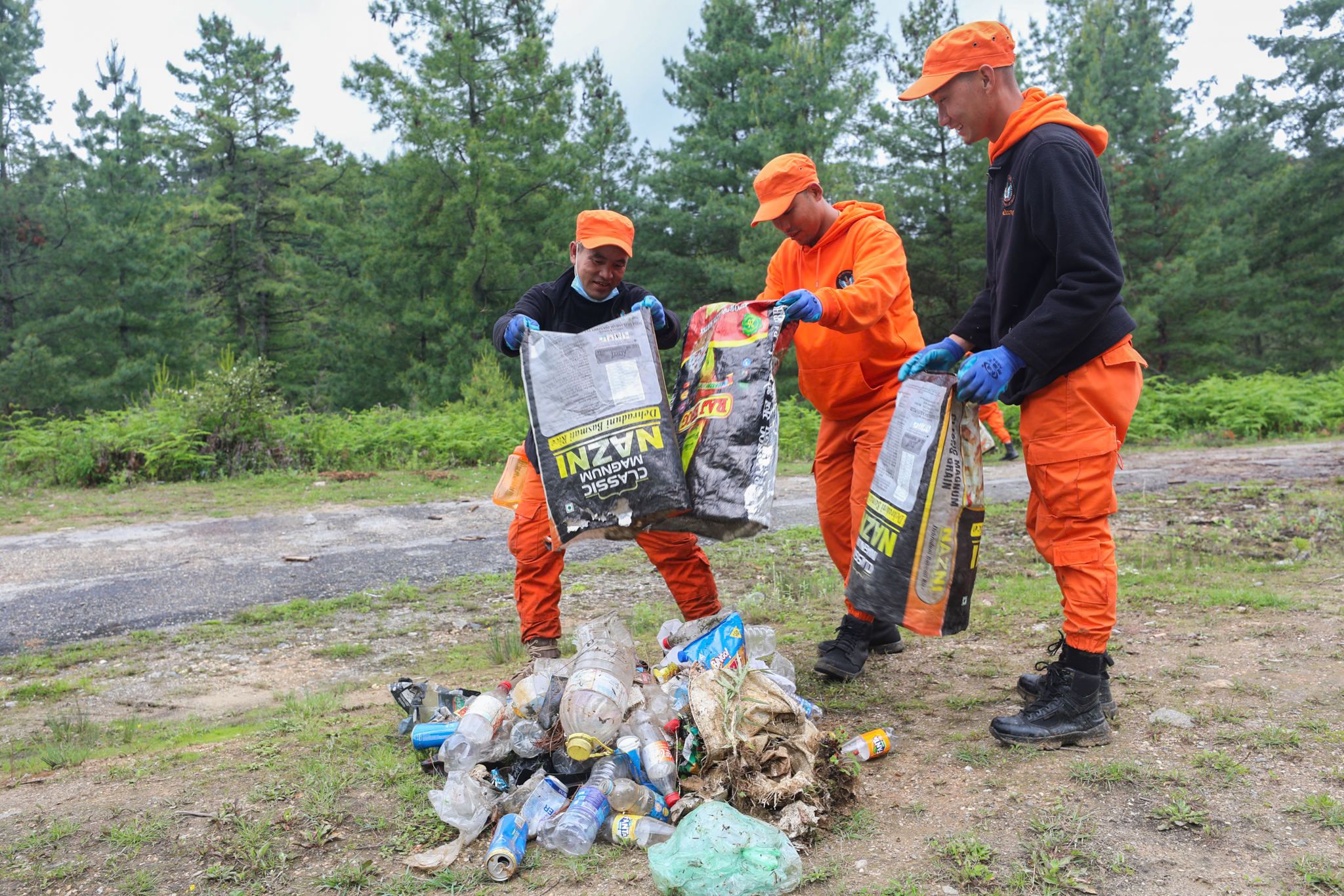 Three people in orange safety gear collect plastic bottles and trash in a forest clearing.