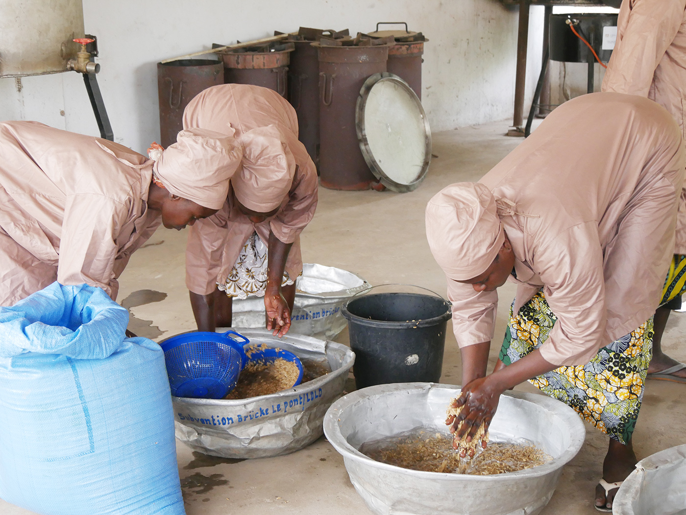 Photo of three people in pink outfits processing food in a rustic kitchen with large bowls.