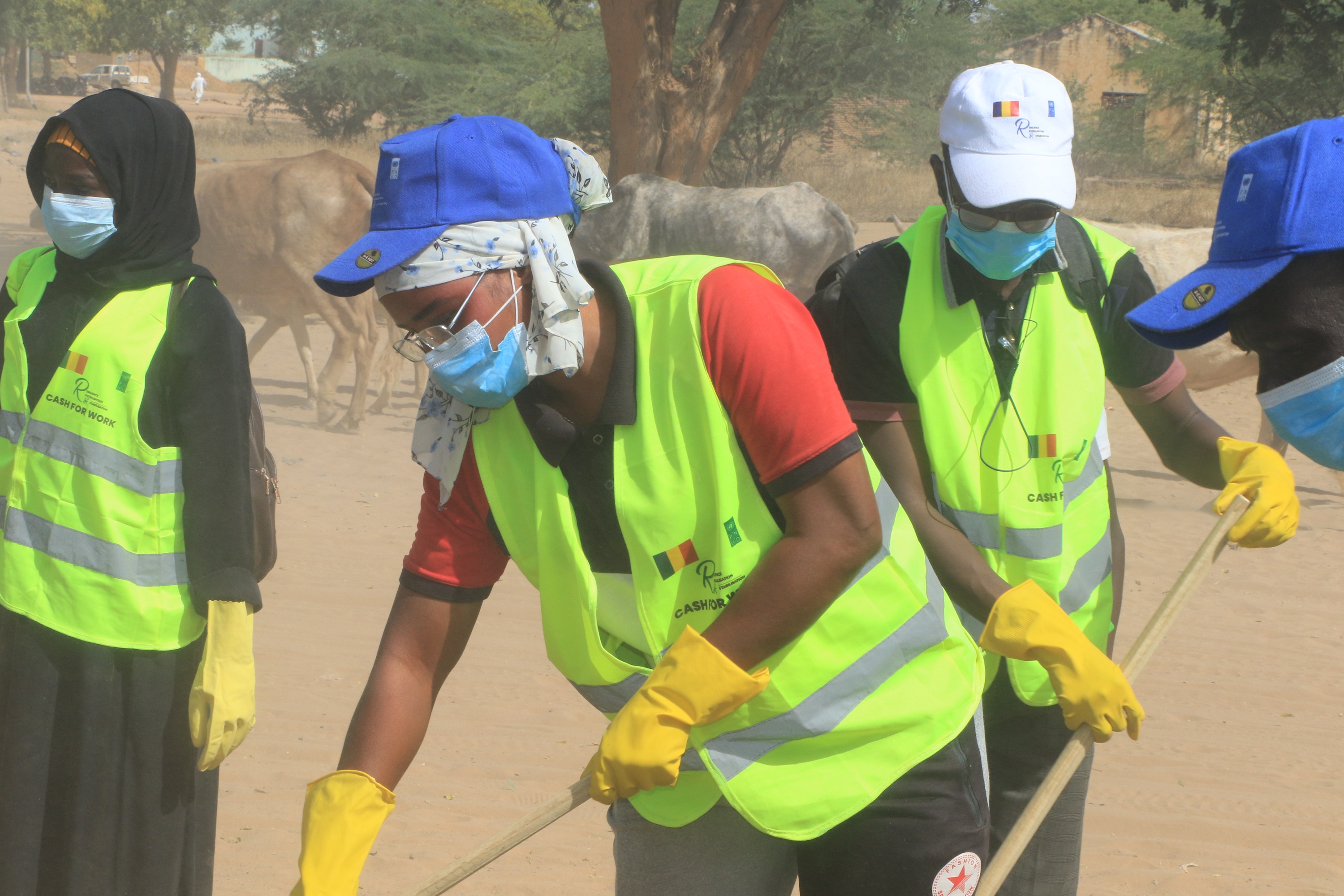 Group of volunteers in neon safety vests and masks picking up litter in a sandy outdoor area.
