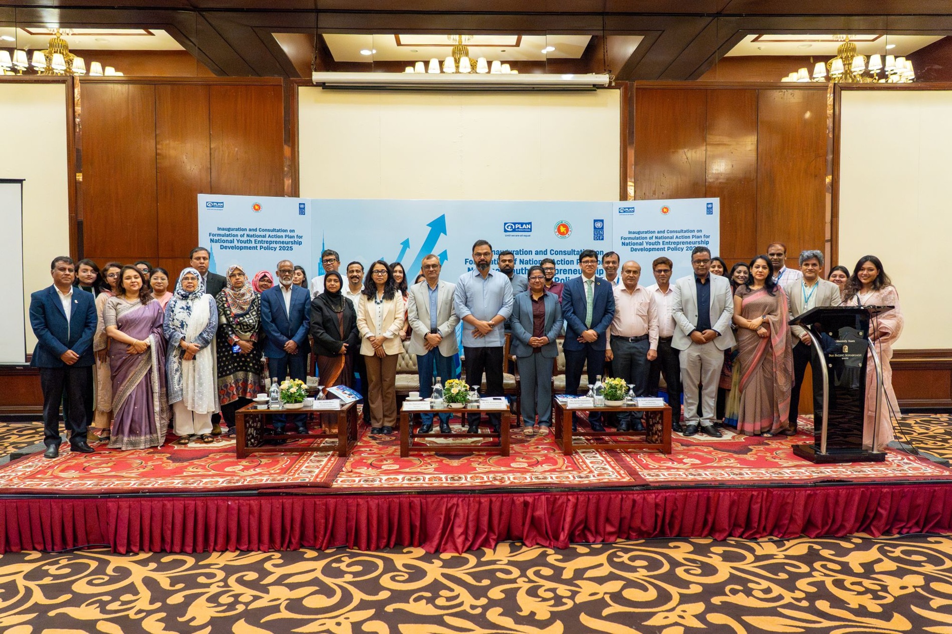 Group of professionals in formal attire posing on stage at a conference in a hotel ballroom.