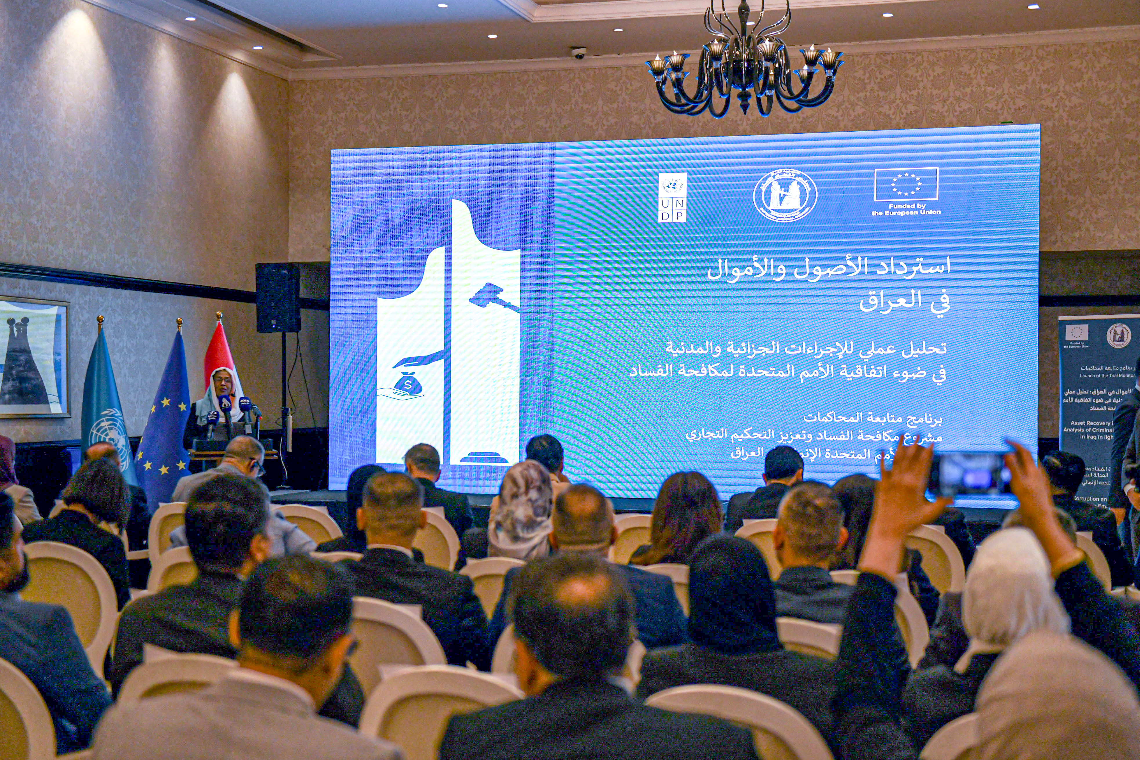 Audience seated in a conference hall watching a large blue presentation screen with logos.