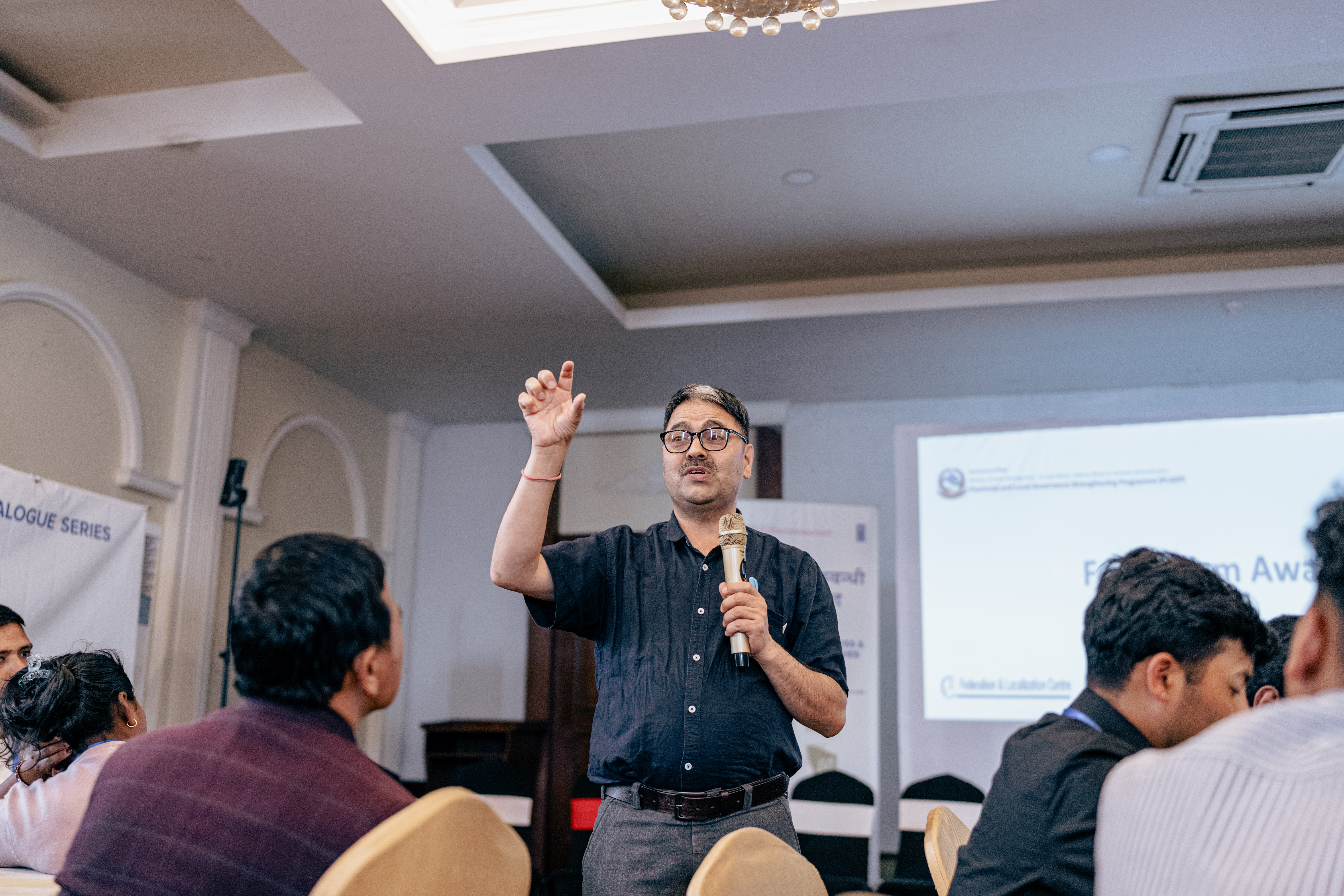 Speaker wearing glasses presents to an audience in a conference room, raising his hand.