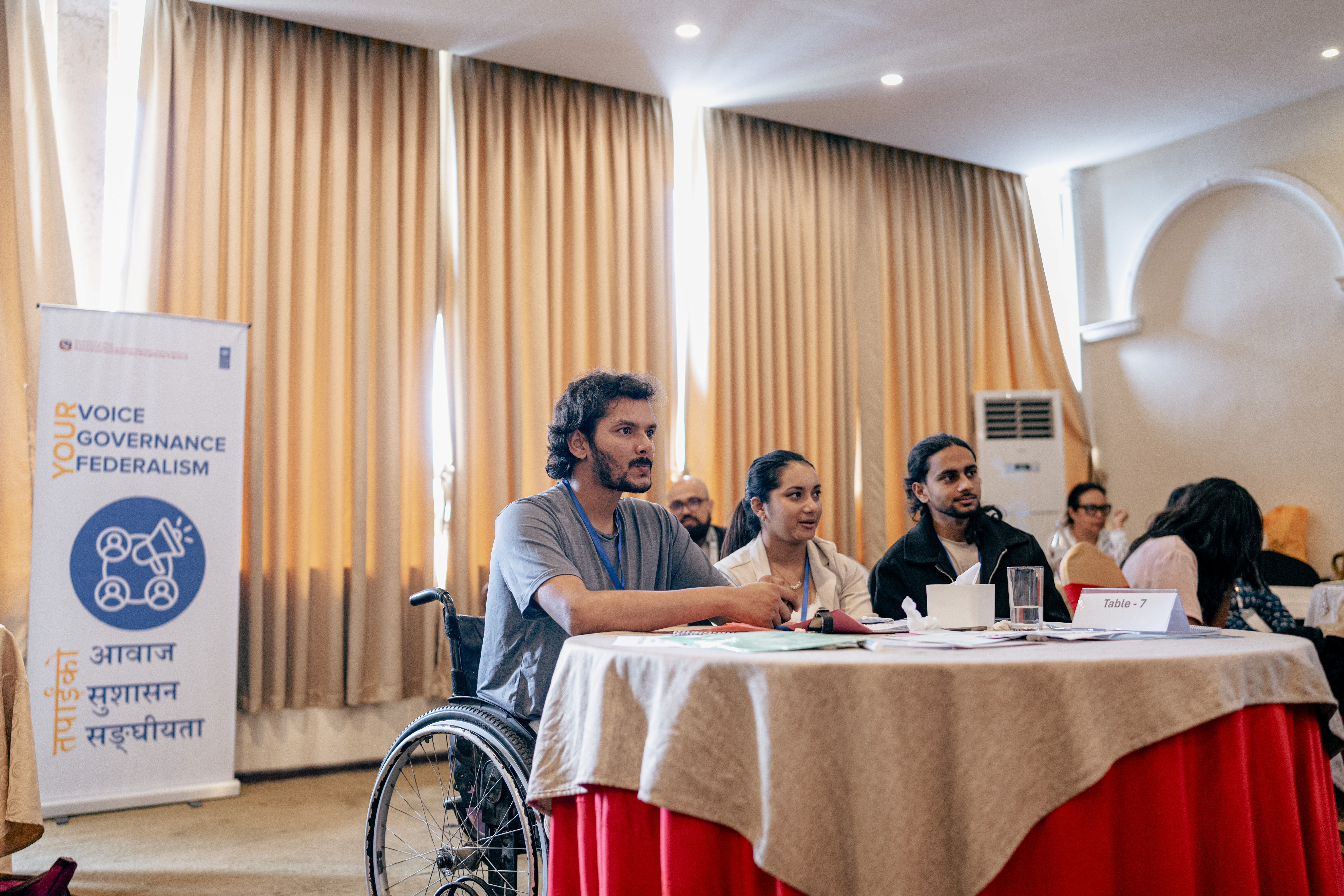 Panel on stage; a wheelchair user sits at a long table with other speakers, beige curtains behind.