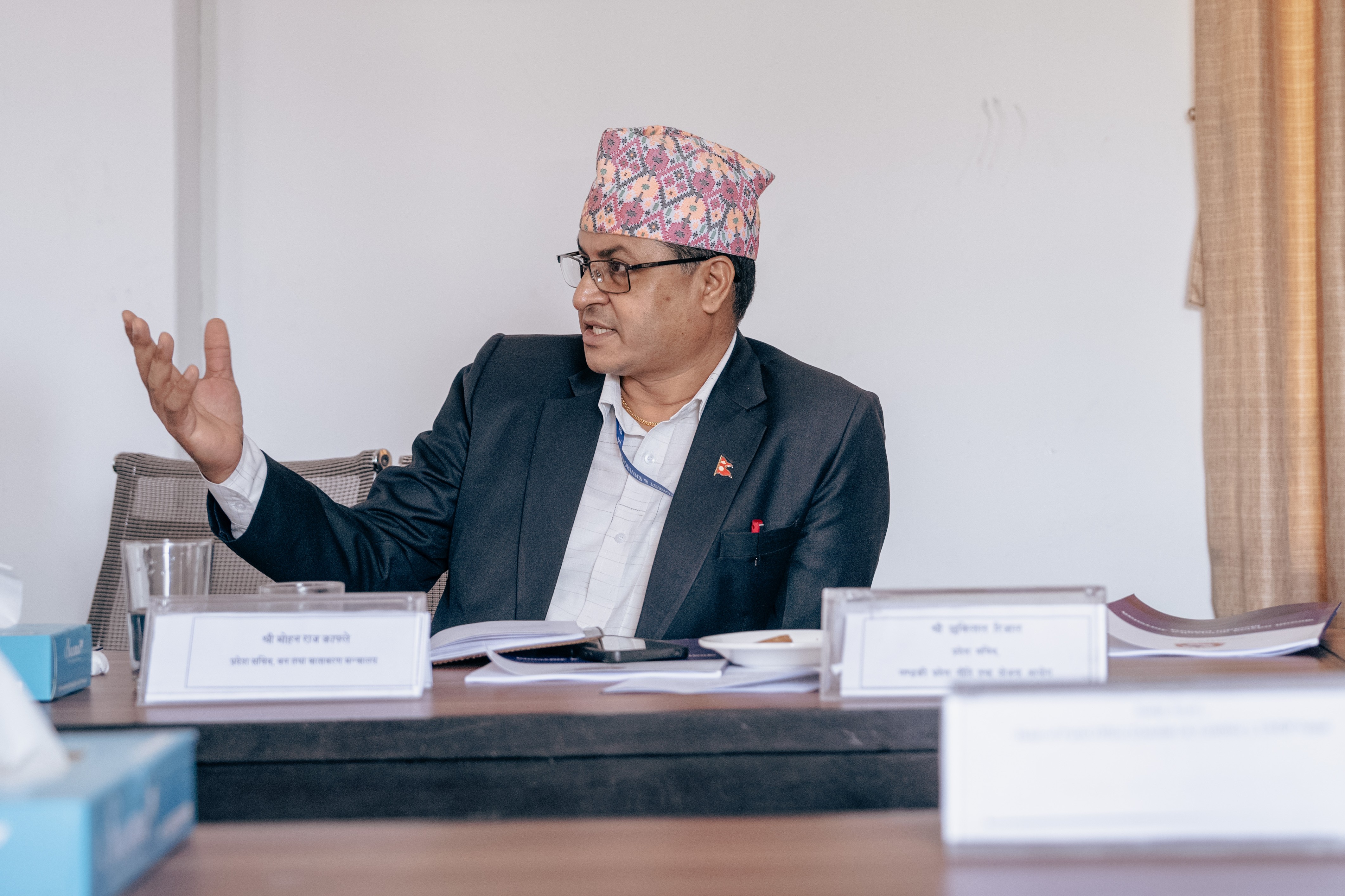 Man in suit and patterned cap gestures at a desk during a formal meeting.