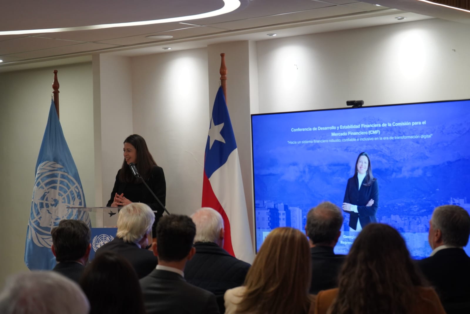 Conference room with speaker at podium, UN and Chile flags, audience watching.