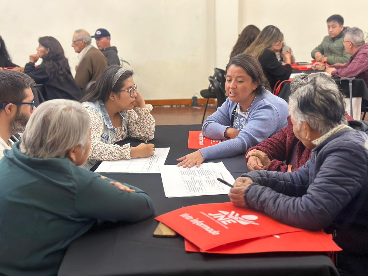 Diverse group of adults seated around a classroom table, discussing papers; red bags nearby.