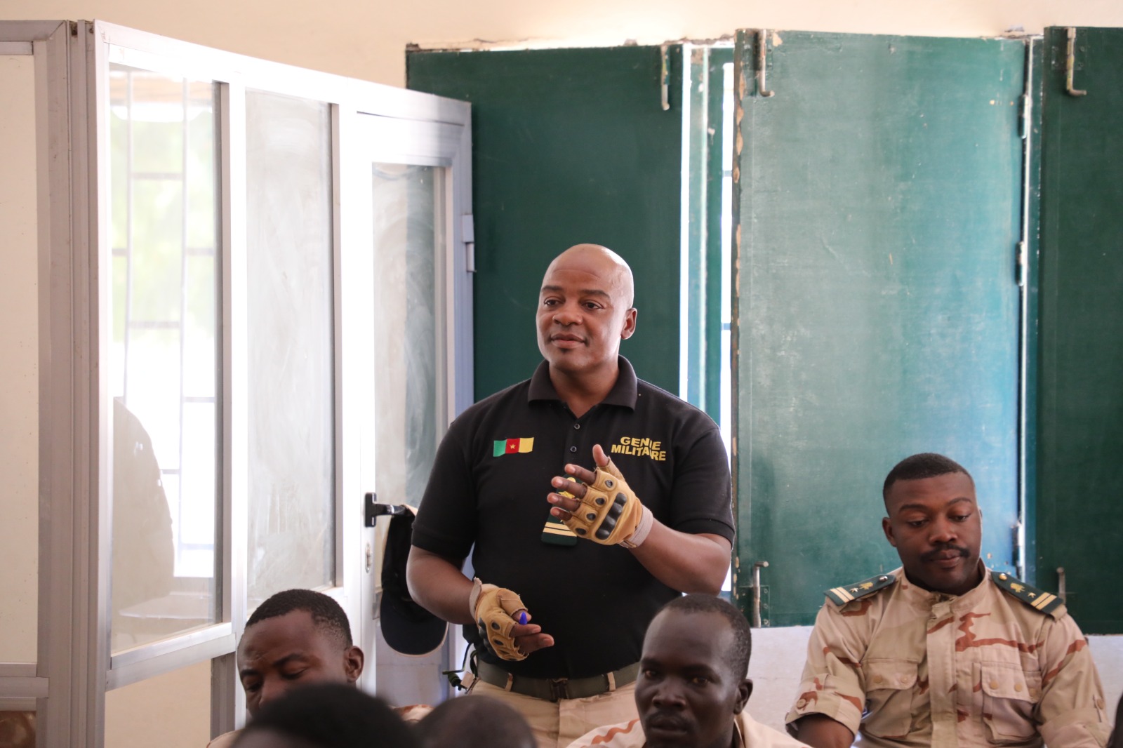 Photograph: bald man in a black shirt speaks to a group in a classroom, holding a Rubik's cube.