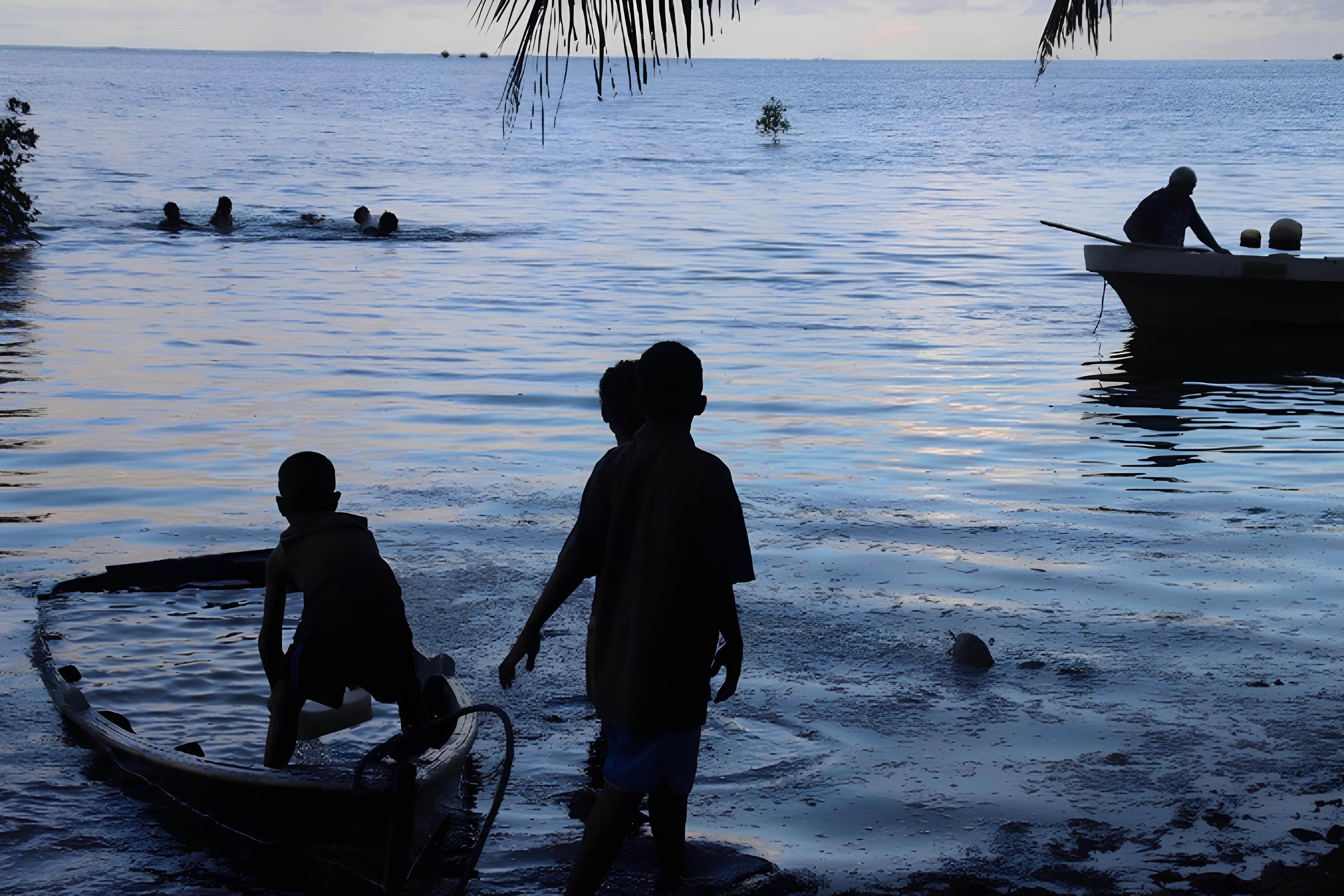 Silhouetted children stand by small boats on a beach at dusk, calm sea and palm trees.