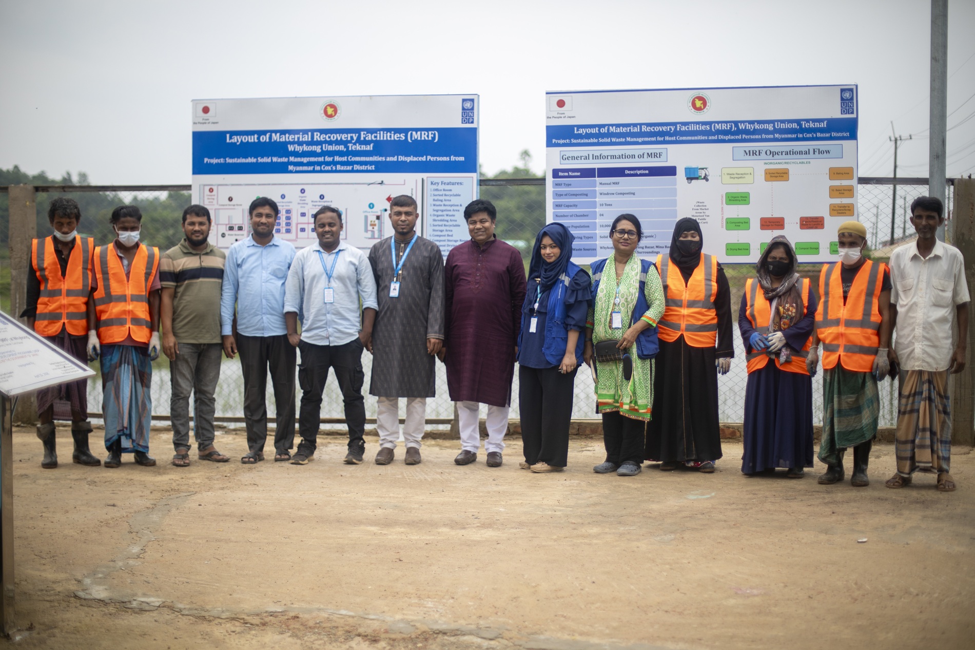 Group of people in orange safety vests and formal wear posing outdoors in front of boards.