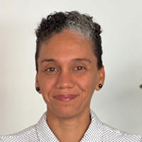 Person with curly hair and earrings wearing a light checkered shirt, pale wall background.