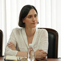 Seated woman at a table with a notebook and water bottle, in a bright room with vertical blinds.