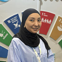 Person in hijab and light blue shirt with lanyard, standing before colorful numbered panels.