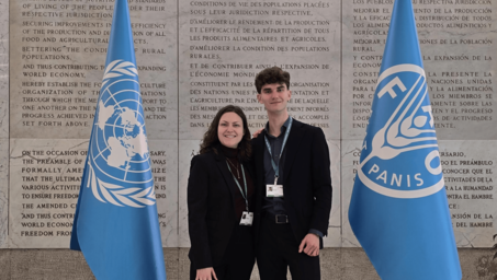 Two young adults pose together in front of UN flags and a stone wall with inscriptions.