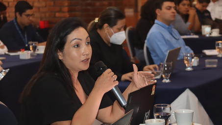A woman speaks into a microphone during a conference, with attendees engaged in the background.