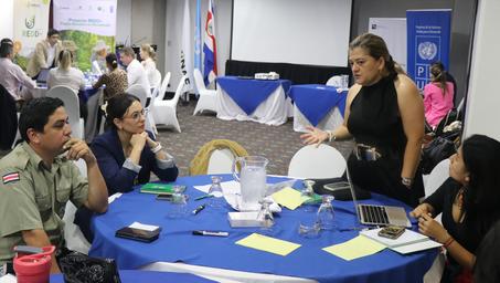 A group discusses at a round table, with a speaker gesturing, and others listening attentively.