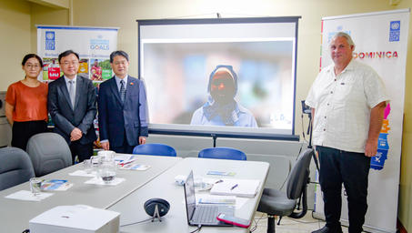 A group of four people stands in a conference room with a video screen displaying a speaker.