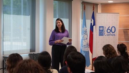 Photograph: Female presenter in purple top speaks to an audience in a conference room.