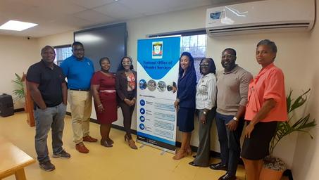 Eight people pose beside a blue-and-white banner with logos in a bright room.