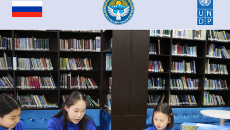 Photograph: three children reading at a library table, shelves behind, flags and logos overhead.