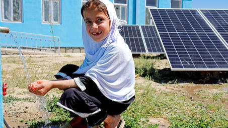 Person crouching beside solar panels in a sunny yard; blue building in background.