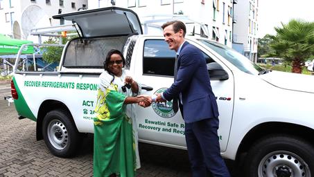 A man in a dark suit hands a key to a woman in a green sari beside a white branded van.