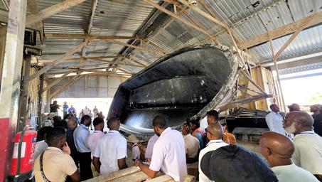 Crowd of people gathered around a large black boat hull under a wooden roof in a workshop.