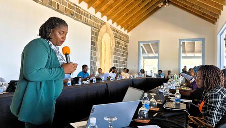 Speaker in teal jacket addresses attendees at a long conference table in a wood-beamed room.