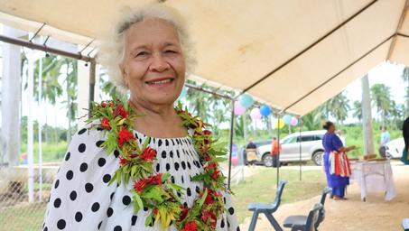 Person with blurred face wearing white polka-dot dress and a colorful lei under canopy outdoors.
