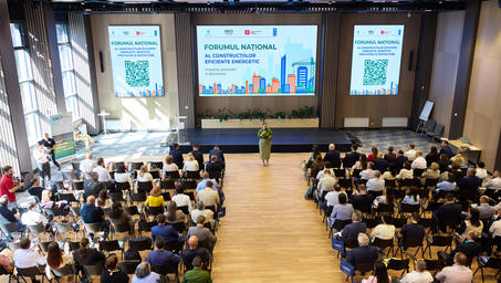 Photograph of a large auditorium with an audience watching a speaker at a podium and two screens.