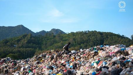 Photograph of a person atop a massive garbage dump, hills in distance and blue sky.