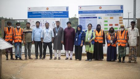 Group of people in orange safety vests and formal wear posing outdoors in front of boards.