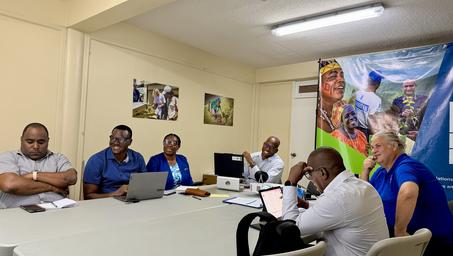 People sit around a rectangular table in a beige meeting room with posters and a colorful banner.