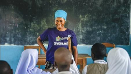 Female instructor in blue hijab and purple shirt addressing seated children in a classroom.