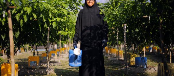 A woman in her 50s, wearing a black headscarf and dress standing in her farm with a watering can.