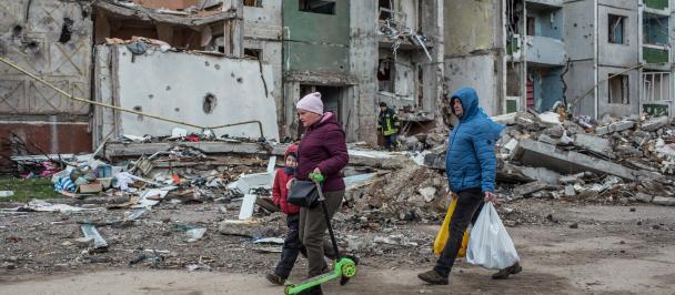 A family walks down a street with damaged buildings in the background.
